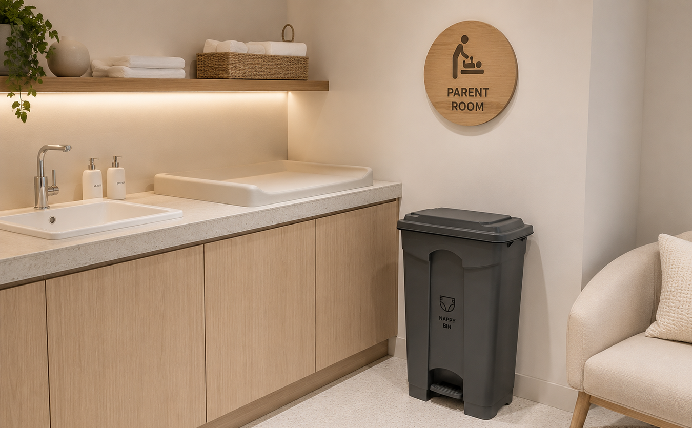 A clean, modern diaper changing room corner featuring light wood cabinets, a double sink, a changing pad, a dark gray trash can, a round wooden 'PARENT ROOM' sign on the wall, and a cream-colored armchair.